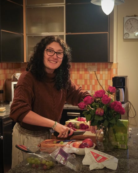 Woman with dark curly hair and glasses in maroon jumper cuts food on a chopping board on kitchen counter strewn with grapes, next to vase of dark pink flowers.