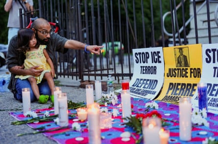 man kneels on ground with young girl and points to signs reading ‘stop ICE terror’ and ‘justice for Pierre’ at vigil with candles and flowers