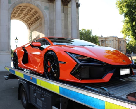A seized orange sports car sits loaded on a flatbed tow truck near a stone archway